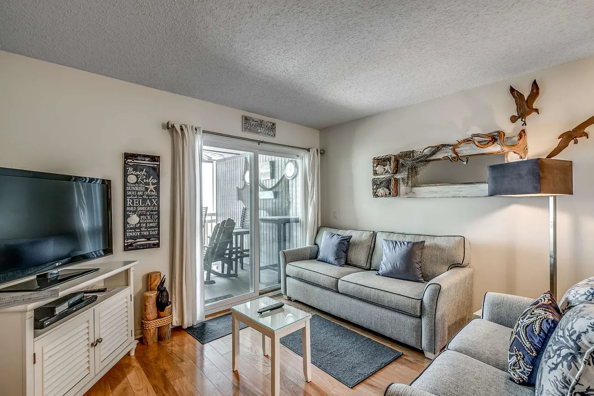 A sunlit living room features a comfortable sofa, television, and a sliding glass door that leads out to a balcony with ocean views. The space is cozy, inviting, and highlights the seamless transition from indoor relaxation to outdoor vistas.
