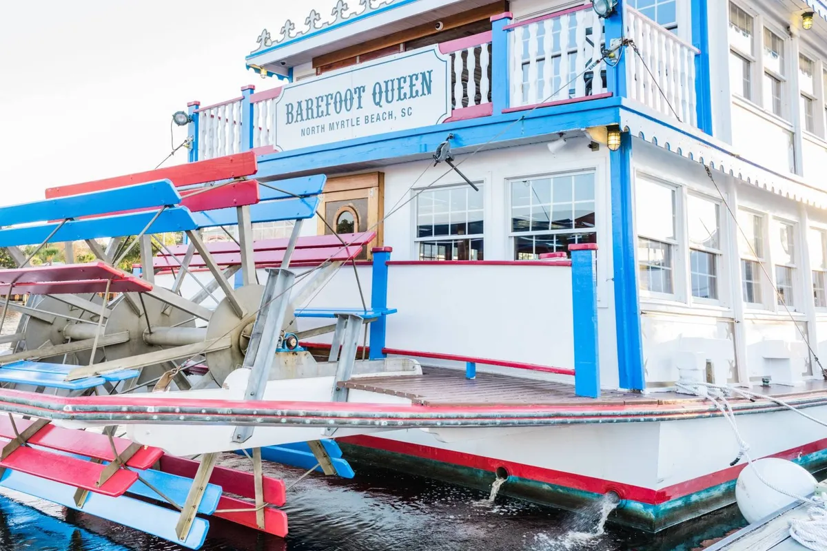A vibrant scene of the Barefoot Queen Riverboat, with its paddlewheel and colorful exterior, docked by the water. Guests can be seen enjoying a festive atmosphere on deck, hinting at live music and waterfront dining experiences.