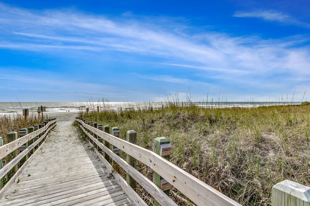 This image captures a wooden walkway leading through dune grass towards a gorgeous ocean view under a blue sky. The scene is tranquil and inviting, suggesting easy access to the beach and opportunities for sunrise walks or playful days by the shore.