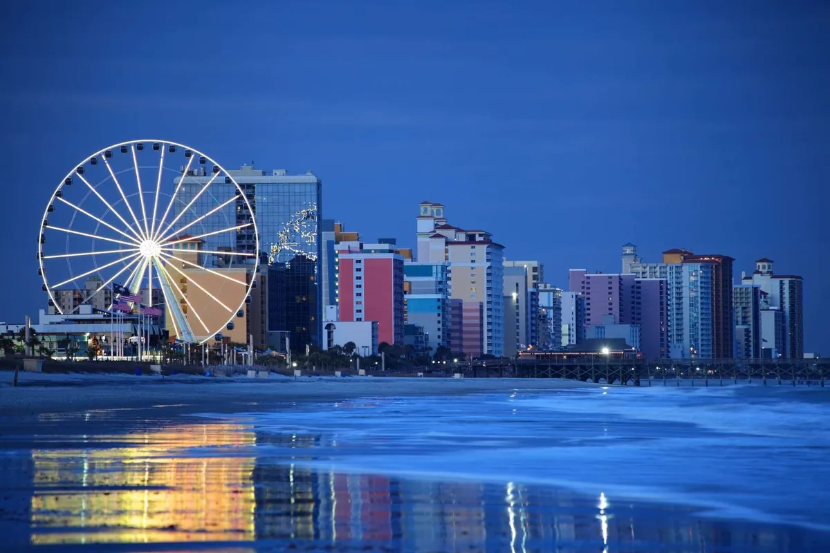 A lively exterior shot featuring a ferris wheel, colorful oceanfront buildings, and shimmering reflections on the water. The festive atmosphere hints at fun amusement rides, family outings, and nights spent exploring the local area.