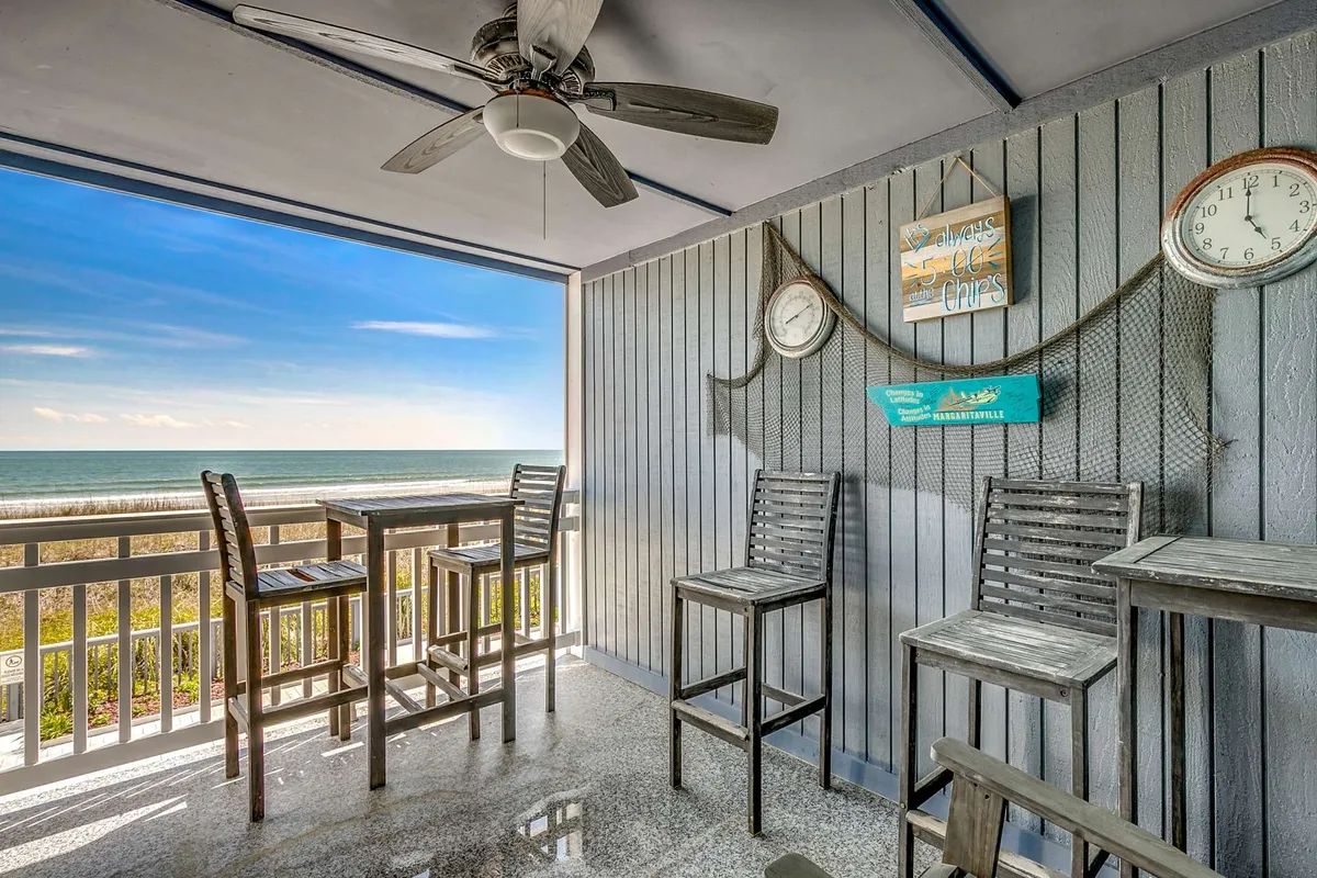 This exterior image shows a relaxed oceanfront setting at Windy Shores I Unit 102, featuring bar stools facing sweeping views of the Atlantic, nautical décor, playful signage, and inviting spots ideal for gathering with friends or family. The scene is perfect for sipping cocktails at sunset or enjoying your morning coffee while listening to the waves.