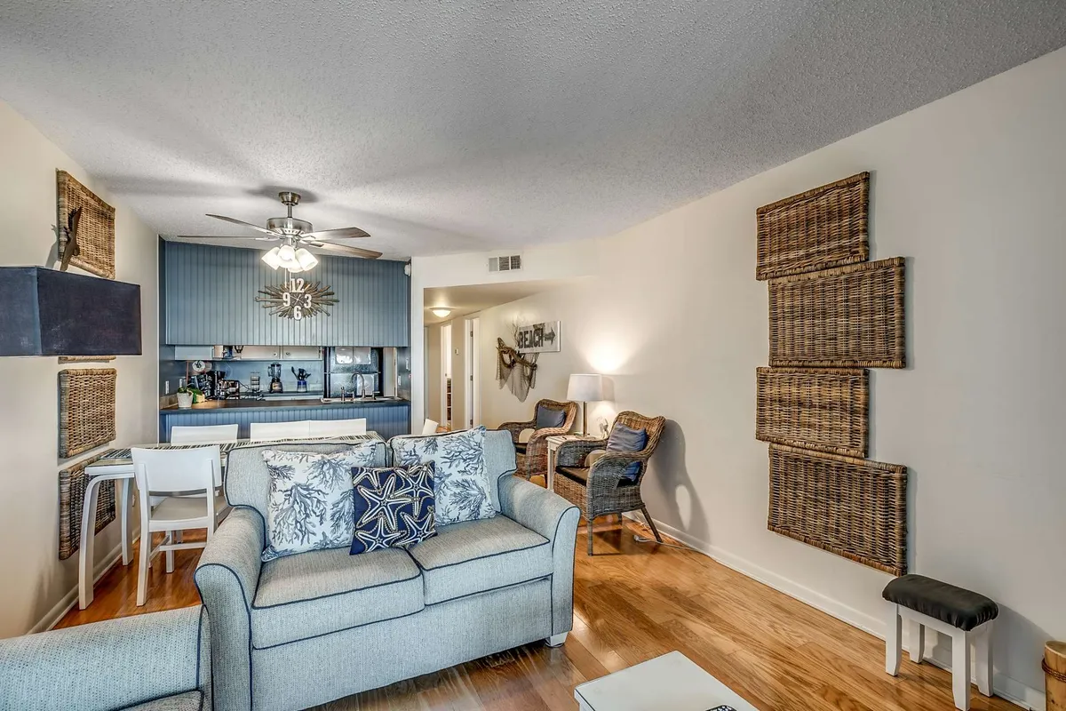 An inviting living space with a plush sofa decorated with accent pillows, a blue feature wall, a dining table with white chairs, woven wall decor, and wood flooring. The setup feels airy and comfortable, encouraging relaxation and socializing.