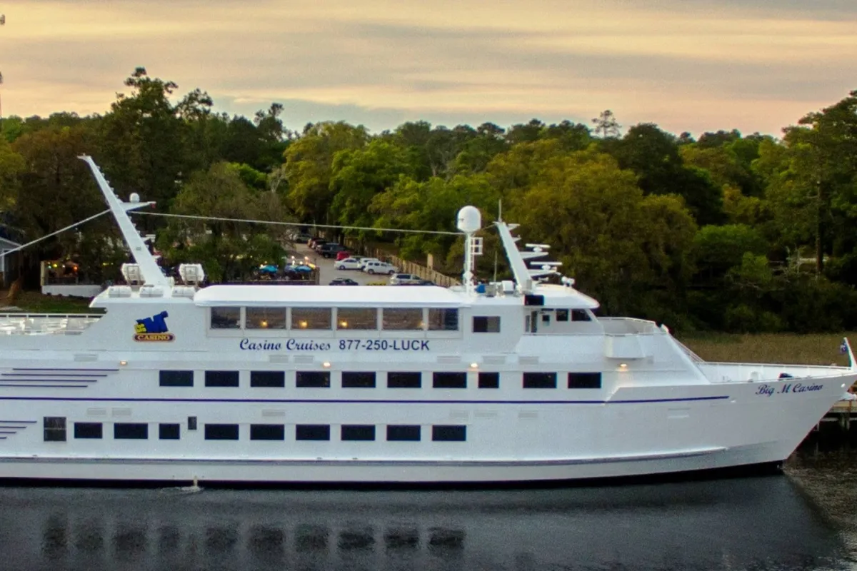A sleek, luxurious yacht is docked amidst lush green trees, with clear casino branding visible. The yacht is set up for dining and entertainment, inviting guests to experience river cruising, dining under the stars, and the excitement of casino gaming in a vibrant yet natural setting.