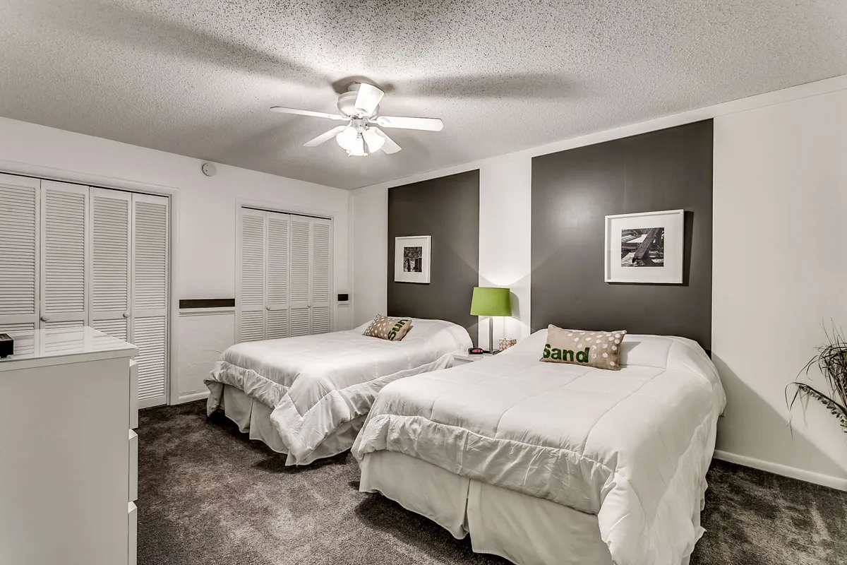 The guest bedroom is depicted with two full beds dressed in crisp white linens, accented by a green lamp, decorative 'sand' pillows, and framed wall art. The room feels peaceful and inviting, designed for rest or casual morning chats.