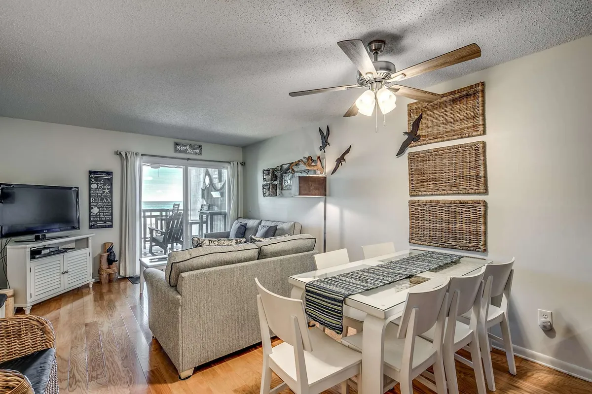 A cozy open-plan living and dining room featuring a plush sofa, a large TV, a modern dining table with chairs, tasteful wall art, and expansive glass doors that open up to a balcony overlooking the ocean. Natural light floods the space, highlighting both the gathering areas and the seamless transition to the outdoors.