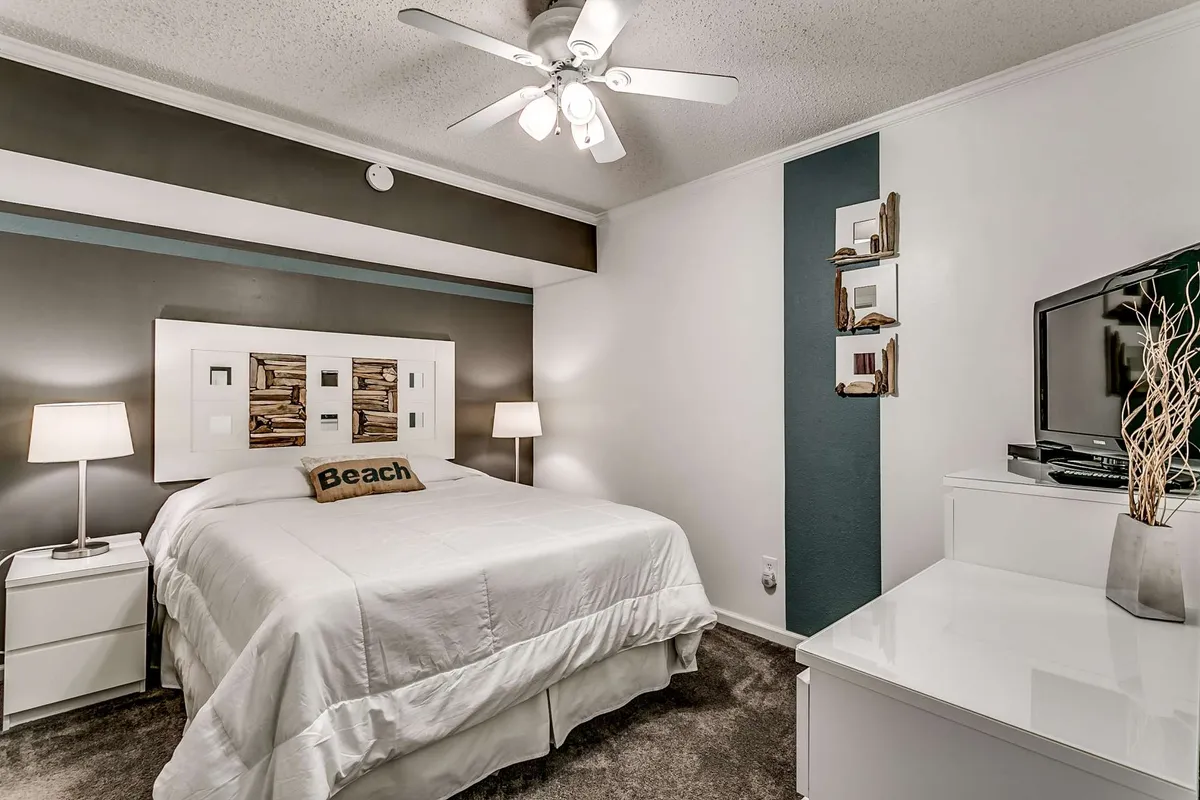 This image features a serene bedroom with a stylish modern headboard, a bed adorned with a beach-themed pillow, a wall-mounted TV, and shelving with decorative items. Soft natural light filters in, creating a tranquil and cozy environment ideal for unwinding.