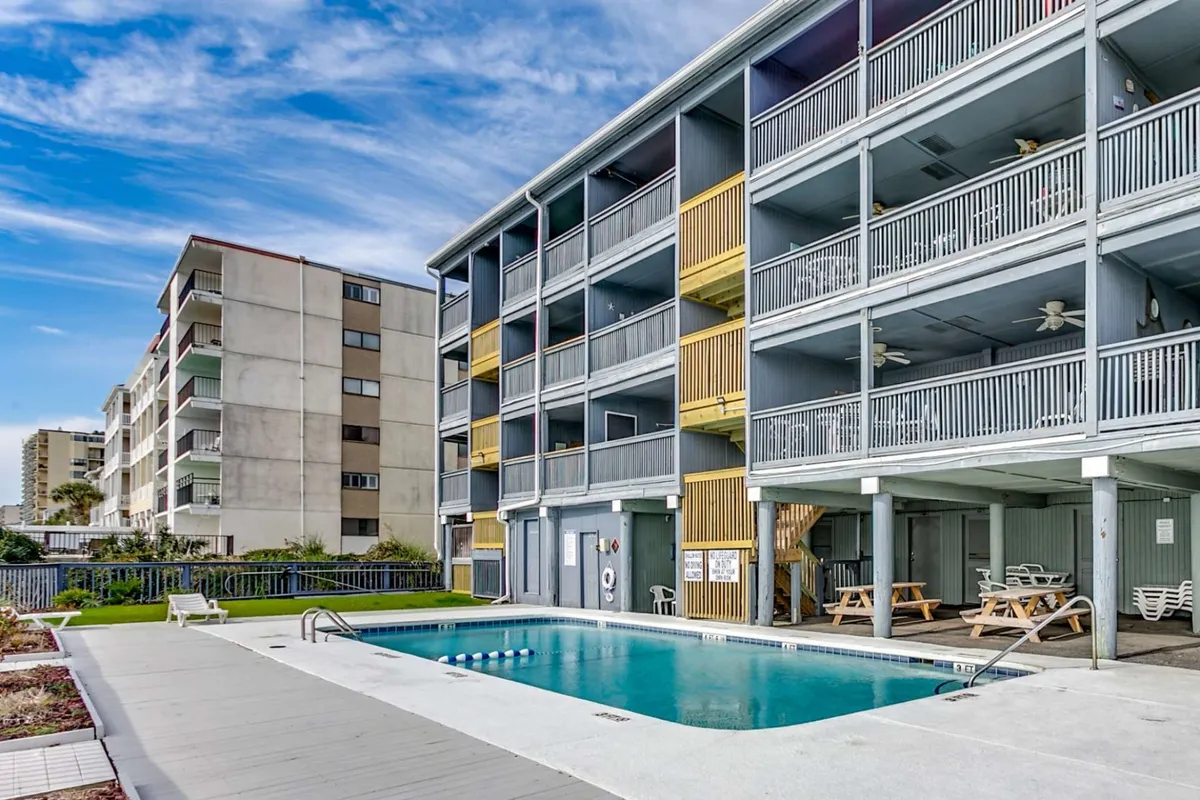 A vibrant exterior scene featuring the communal swimming pool, surrounded by lounge chairs and balconies overlooking the area. The pool is inviting, with the building's balconies suggesting easy access for guests. The atmosphere is lively and social, hinting at group relaxation and fun under sunny skies.